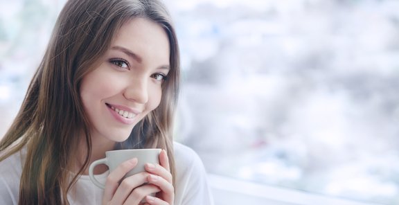 Schöne lächelnde junge Frau mit natürlichem Make-up und langen Wimpern hält eine Tasse mit heißem Kaffee oder Tee. Wintersaison. 